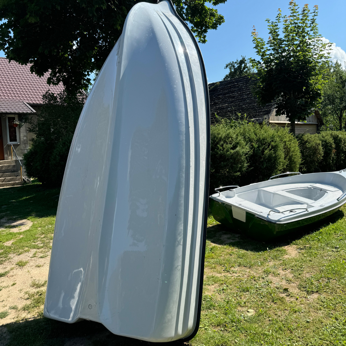 Biała łódź postawiona pionowo na rufie na trawniku, eksponująca profil dna kadłuba; obok leży druga łódź.